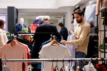 Holiday discounts. Young millennial guy choosing t-shirt in outlet store, looking through...