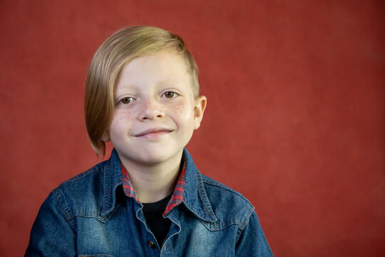 Niño rubio sonriente de frente con camisa azul y fondo rojo