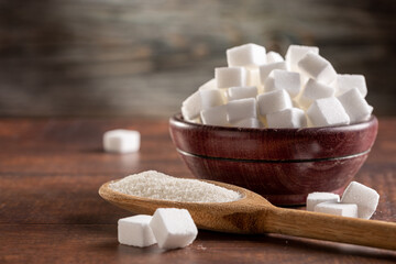 White sugar cubes in wooden bowl and granulated sugar on wooden spoon.