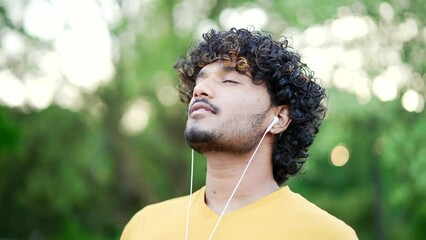Close up. Happy young adult man in headphones relaxing with closed eyes standing in urban city park. Smiling handsome male in a t-shirt takes deep breaths and enjoys being outdoors, listening to music - Powered by Adobe