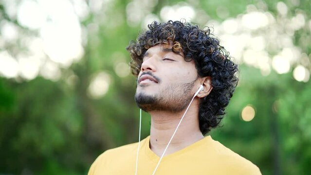Close up. Happy young adult man in headphones relaxing with closed eyes standing in urban city park. Smiling handsome male in a t-shirt takes deep breaths and enjoys being outdoors, listening to music