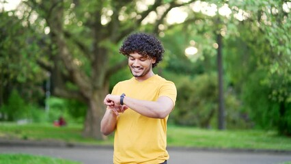 Happy young runner checking workout results on smart watch at urban city park. Fit handsome man in a T-shirt smiles and rejoices at his success looking at a smartwatch bracelet, showing a yes gesture 