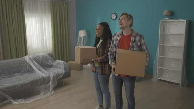 Young couple with cardboard boxes in their hands in a new apartment. A man and an African American woman are moving into a new flat, they are pleased to inspect their new home. HDR BT2020 HLG Material