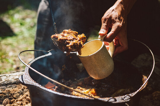 Hands ladle a rustic stew into a much from a cast iron pot in an outdoor setting
