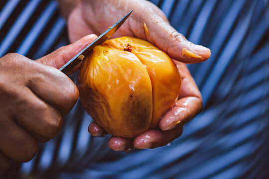 Wet hands peel a peach with a knife in front of blue striped cloth