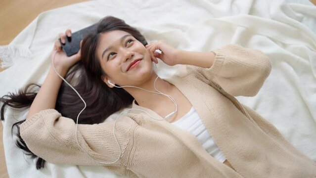 Young asian woman listening to music on the floor in living room at home. Happy asia female using mobile smartphone, wearing headset and lying down on carpet