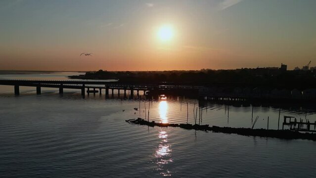 An aerial view over the Jamaica Bay, facing the sun at sunrise. A train crosses the bridge as the sun reflects onto the calm waters. The drone camera truck left and pan right over the water.