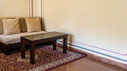 A seating area in the living room. On the red patterned carpet there is a sofa with pillows, a wooden coffee table. Beige walls, wooden floor.