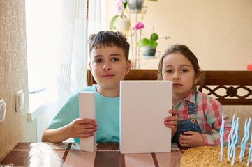 Authentic portrait of a teen boy and his younger sister holding white mockup box , sitting at table and celebrating happy life event. White gift boxes with copy space for advertising text - Powered by Adobe
