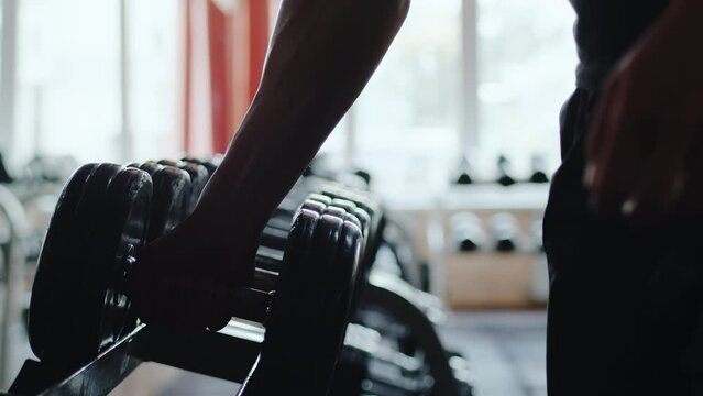 Athlete approaches the rack with sports equipment and takes a heavy dumbbell close up. Sportsman preparing to power exercise during workout in an indoor gym