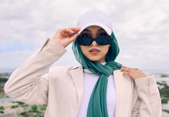 Portrait, fashion or sunglasses with a saudi woman outdoor in a cap and scarf for contemporary style. Islam, face and hijab with a trendy young muslim or arab person posing outside in modern clothes