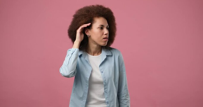 Cute curious african american woman keeps hand near ear tries to overhear something very interesting listens somebodys secret or collecting rumors isolated on pink studio background