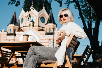 Portrait of glamorous young woman in sunglasses sitting at street cafe table on sunny day, looking...