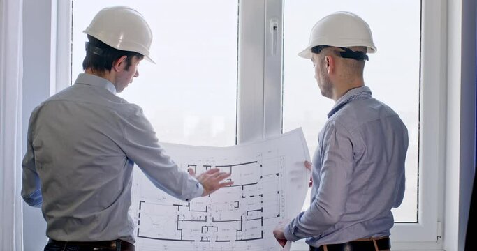 Engineers in hardhats and office clothing have discussion about flat drawing while collate plan with room. Employees stand near window talking