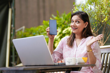 Indian woman showing smartphone screen while using laptop.