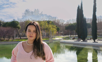 portrait headshot of a pretty woman in nice make up looking at camera with neutral expression and a lake of a park in the background