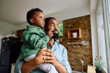 Black father holds his small son as they look out the window together at home.