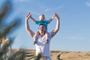 Dad and his little son are having fun walking in a field with ripe wheat. The child is sitting on the shoulders of the father.