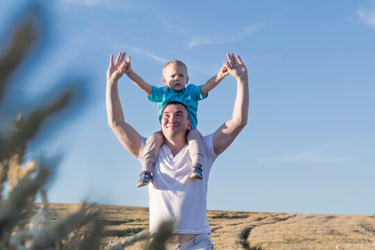 Dad And His Little Son Are Having Fun Walking In A Field With Ripe Wheat. The Child Is Sitting On The Shoulders Of The Father.