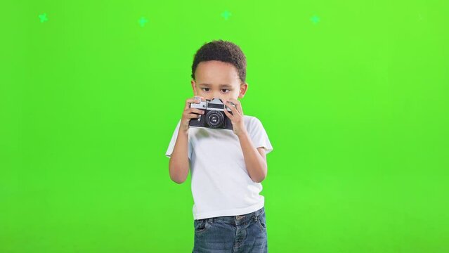 Fun little kid boy in white t-shirt holding retro vintage photo camera, doing photo shot, isolated on green background. Front view of african american child taking photo in motion. Lifestyle concept.