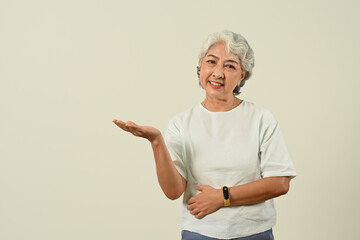 Cheerful grey haired mature woman standing over beige background presenting at empty space