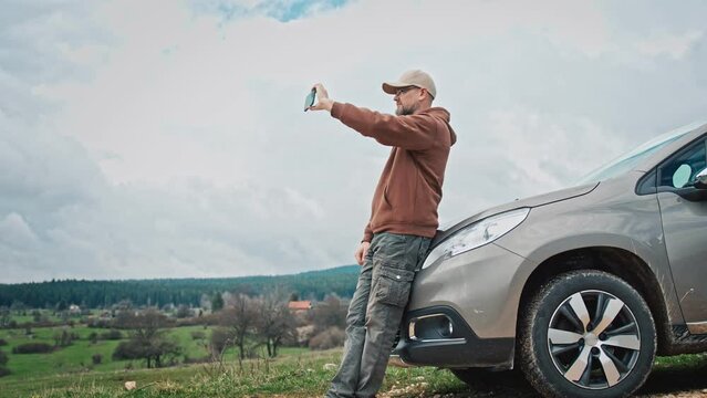 An adult man taking a picture of the view leaning against the hood of his car during a solo car trip.