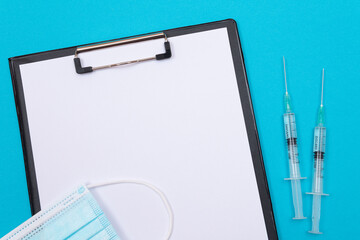 Vaccination, Immunology or Revaccination Concept - Two Medical Syringe Lying on Blue Table in Doctor's Office in a Hospital or Clinic. Black Clipboard with Sheet of Paper - Mock Up with Copy Space