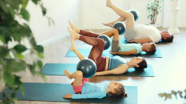 At home, mom, dad, son and daughter engaged in physical education Pilates with softball. Middle-aged parents with children lie on backs on koremat and raise legs bent at the knees
