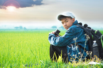 Asian boy in plaid shirt and cap sitting on paddy field in the morning and holding binoculars in  hands to watch birds which flying on the sky and eating fish in rice paddy field, summer vacation.
