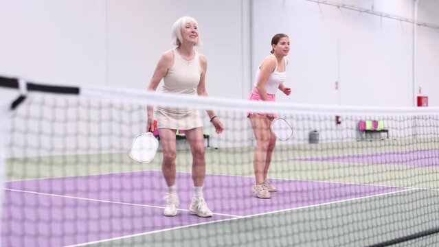 Two athletic women of different ages are playing a game of pickleball on a court inside a sports facility