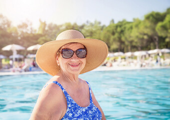 Senior woman (over age of 50) in swimsuit, hat and sunglasses relaxing in resort swimming pool. Happy retired female enjoying summer vacation in hotel. All-inclusive..
