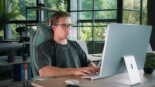 Young caucasian male working at the desktop computer, smiling and nodding. An interesting job, a funny task in a cozy informal office