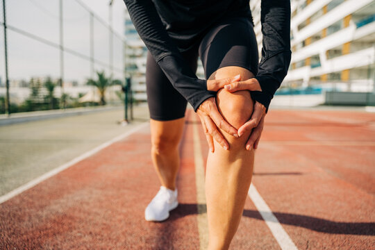 Close Up Of Sport Woman Holding Knee With Hands In Pain After Suffering Injury Running. Runner Woman Sit On Road With Knee Injury And Pain. 