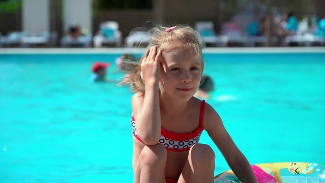 Little girl rests by hotel pool with blue water and inflatable swim ring and smiles. Summer vacation, holiday and travel concept