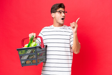 Young caucasian man man holding a shopping basket full of food isolated on red background intending...