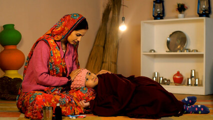 A sick young girl wearing a hat and covered with a shawl resting on her mother's lap - cold and...