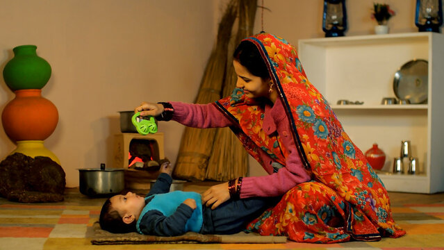 A beautiful young lady of an Indian village playing with a cute baby girl at home - parenting  single mother  village home. Village scene of a rural housewife in a printed Saree spending quality ti...