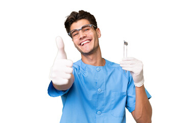 Young dentist man holding tools over isolated background with thumbs up because something good has...