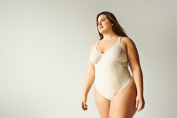 happy plus size woman in beige bodysuit posing while standing in studio on grey background, body positive, figure type, self-esteem, smiling while looking away