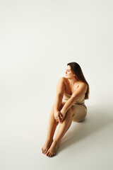 full length of barefoot and confident woman with plus size body in strapless top with bare shoulders and underwear posing while sitting in studio on grey background, body positive, figure type