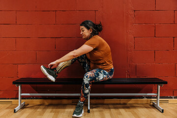 Side view of athlete with disability wearing shoe while sitting on bench near red wall at sports court