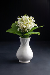 On the table in a white vase is a bouquet of lilies of the valley. On a black background.