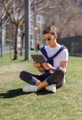 Fototapeta premium a young man is sitting with a tablet in his hand in a park, concept of remote work, online communication, using social networks, studying or working outdoors