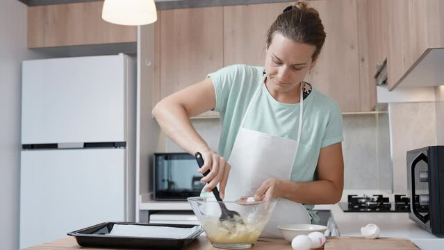 In a bright kitchen interior, a young woman is stirring the choux pastry with a spatula in slow motion.