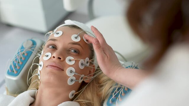 Closeup view from shoulder of unrecognizable physiotherapist consulting young woman client during facial myostimulation procedure in beauty clinic. Concept of rejuvenating skin, preventing aging.