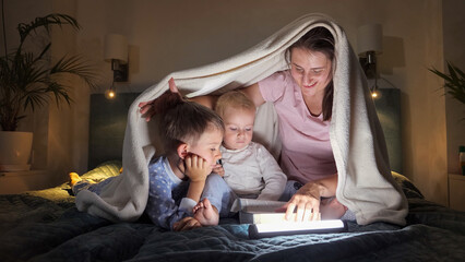 Two boys and mother in pajamas reading bedtime story book in bed at night with flashlights. Family having time together, parenting, happy childhood and entertainment.
