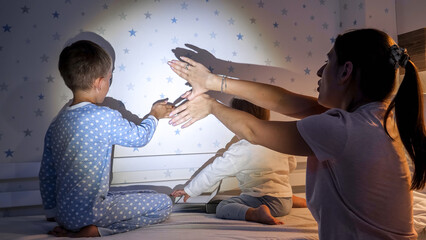 Young mother with kids making theater of shadows on wall in bedroom. Family having time together,...