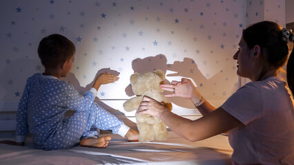 Smiling boy with mother showing theater of shadows with flashlight on the wall. Family having time together, parenting, happy childhood and entertainment.