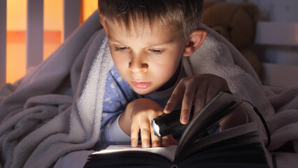 Closeup of boy holding flashlight reading book in bed. Children education, development, secrecy, privacy, reading books. - obrazy, fototapety, plakaty