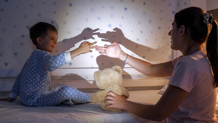 Cute smiling boy with mother playing with shadows from their hands on wall from bright flashlight light - obrazy, fototapety, plakaty